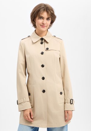 Frau trägt einen beigen Trenchcoat mit schwarzen Knöpfen und Gürtel an den Manschetten, steht vor einem einfarbigen hellen Hintergrund.