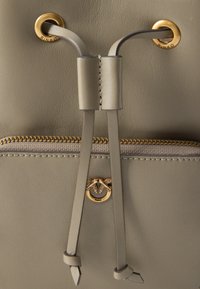 Mochila de couro cinza com um fecho central tipo cordão, hardware em tom dourado e um bolso frontal com ziper que apresenta um logotipo decorativo.