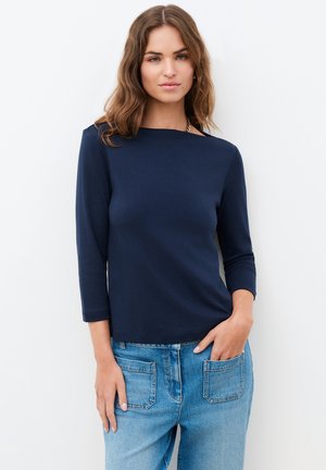 Nuori nainen, jolla on aaltoilevat ruskeat hiukset, pukeutunut laivastonsiniseen pitkähihnaiseen yläosaan ja vaaleansinisiin farkkuihin, seisoo käsi yhdessä taskussa.