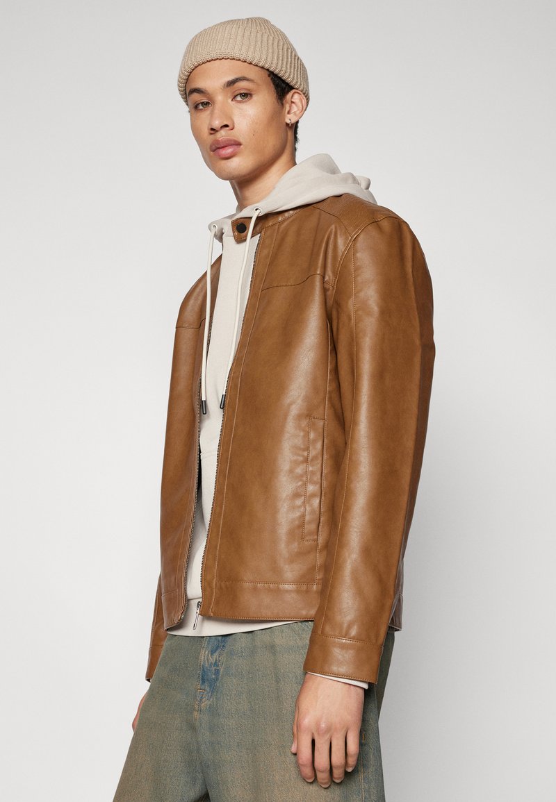 Chaqueta de cuero marrón con una textura suave, que cuenta con un cierre de cremallera y dos bolsillos laterales, llevada sobre un hoodie de color claro.