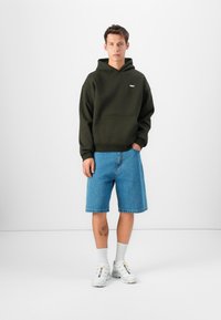 Donkergroene hoodie met een voorzak, gecombineerd met lichtblauwe denim shorts. Witte sneakers en ribbelige witte sokken maken de outfit compleet.