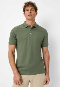 Olive green polo shirt with a collared neckline and short sleeves. Features a blue button at the top and subtle branding on the chest.