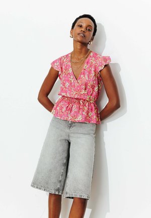 Femme portant un chemisier portefeuille fleuri rose avec manches à volants et un short en jean gris clair longueur genou, debout sur un fond blanc.