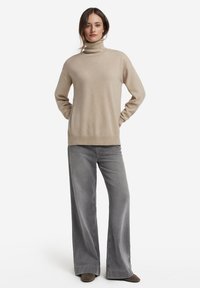 Maglione beige a collo alto con vestibilità rilassata, texture morbida e orlo a coste, abbinato a pantaloni grigi a gamba larga.