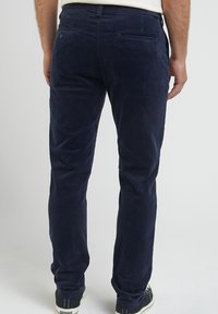 Marinblå kordflöjelbyxor med slim fit. Har bakfickor och en slät yta. Styld med en svart sneaker mot en enfärgad bakgrund.