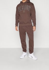 Sweat à capuche marron avec imprimé "NIKECOURT", pantalon de survêtement marron assorti et baskets blanches. Le tissu semble doux avec une coupe décontractée.