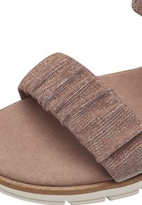 Platte sandaal met een geweven band in bruine en gouden tinten, geplaatst op een glad, lichtbeige voetbed met een stevige witte zool.