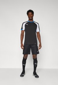 Tricou sport negru cu accente gri și bleumarin, asociat cu pantaloni scurți negri și șosete negre până la genunchi. Logo-ul este afișat proeminent.