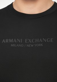 Primo piano di un collo di una maglietta nera con il testo in rilievo "ARMANI EXCHANGE MILANO / NEW YORK" sul petto.