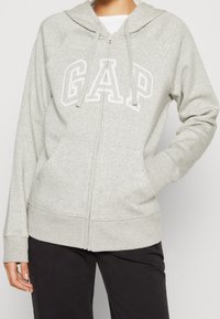 Sudadera gris con cremallera, fabricada con tela suave. Presenta un logo blanco "GAP", cordones ajustables y bolsillos tipo canguro. Puños y dobladillo acanalados.