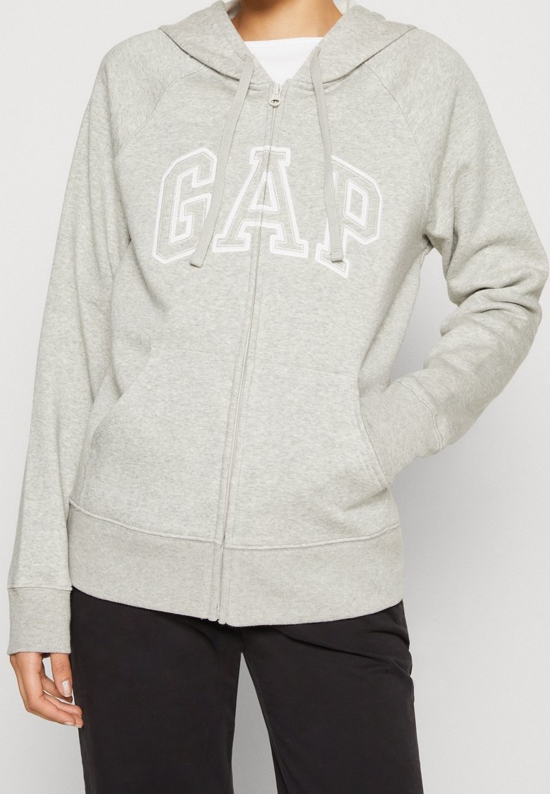 Sudadera gris con cremallera, fabricada con tela suave. Presenta un logo blanco "GAP", cordones ajustables y bolsillos tipo canguro. Puños y dobladillo acanalados.