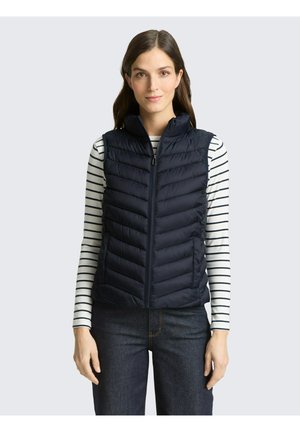 Kobieta ubrana w granatową pikowaną kamizelkę bez rękawów na białą i czarną bluzkę w paski z długim rękawem oraz ciemne dżinsy, stojąca na jasnoszarym tle.