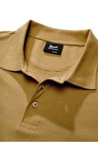 Polo beige de taille moyenne Brandit avec col plié, deux boutons et étiquette de marque visible à l'intérieur de l'encolure.