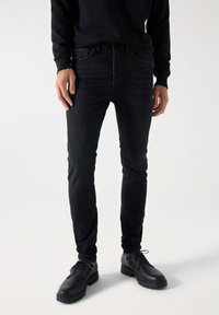 Jeans negras ajustados de mezclilla, con cinco bolsillos y un cierre clásico de botón. Combinados con zapatos negros de cordones.