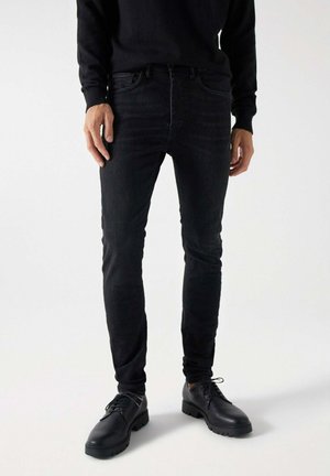 Jeans negras ajustados de mezclilla, con cinco bolsillos y un cierre clásico de botón. Combinados con zapatos negros de cordones.