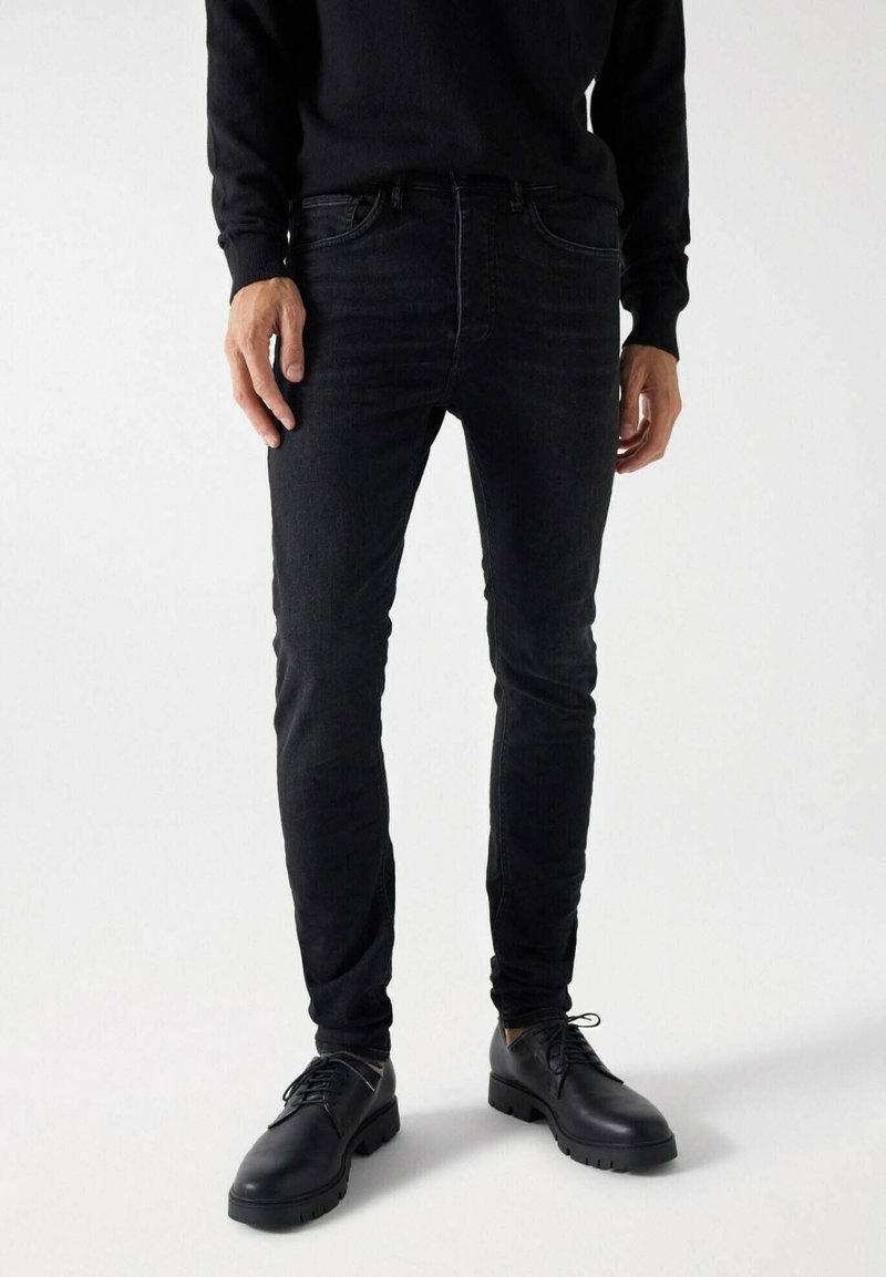 Jeans negras ajustados de mezclilla, con cinco bolsillos y un cierre clásico de botón. Combinados con zapatos negros de cordones.