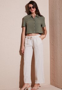 Olivengroene, cropped button-up blouse, gecombineerd met losse, witte linnen broeken. Model met ronde zonnebril, staand tegen een gestructureerde muur.