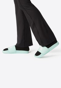 Light green slide sandals with a soft strap, black footbed, and embossed logo. Paired with black athletic pants featuring side seams.