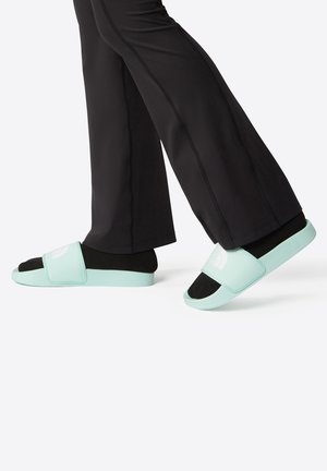 Chanclas de color verde claro con una correa suave, plantilla negra y logo en relieve. Combinadas con pantalones deportivos negros con costuras laterales.