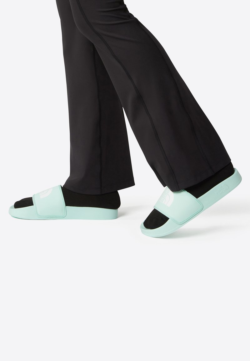 Light green slide sandals with a soft strap, black footbed, and embossed logo. Paired with black athletic pants featuring side seams.