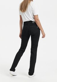 Mujer con cabello largo vistiendo una camiseta blanca, jeans negros ajustados y zapatillas blancas, de pie con una pierna ligeramente doblada.