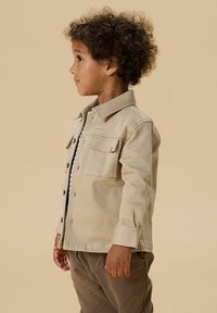 Malé dítě s kudrnatými vlasy, oblečené v béžové bundě na knoflíky a hnědých kalhotách, stojící bokem na jednobarevném béžovém pozadí.