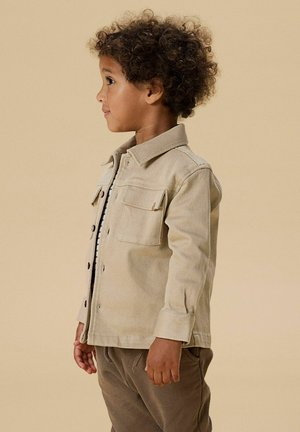 Ungt barn med lockigt hår som bär en beige jacka med knappar och bruna byxor, stående i profil mot en enfärgad beige bakgrund.