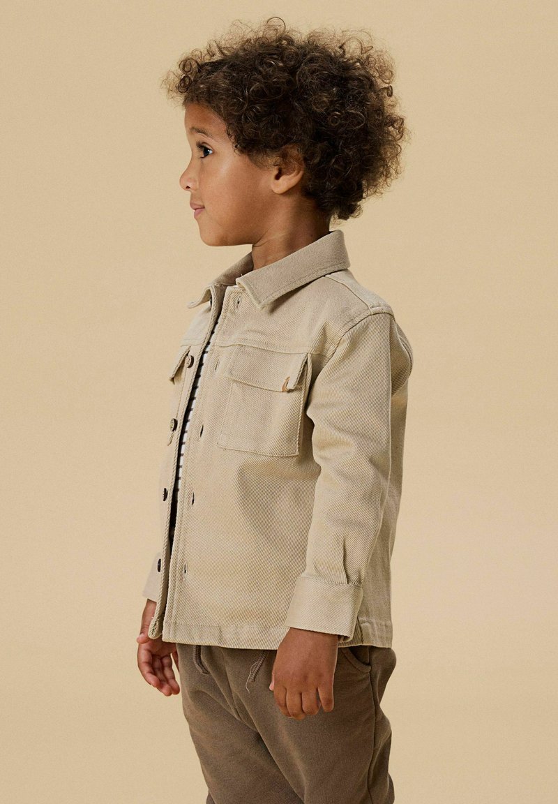 Malé dítě s kudrnatými vlasy, oblečené v béžové bundě na knoflíky a hnědých kalhotách, stojící bokem na jednobarevném béžovém pozadí.