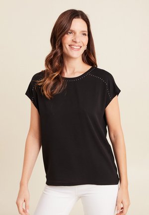 Donna sorridente con lunghi capelli ondulati, che indossa una maglietta nera a maniche corte con piccoli borchie decorative e pantaloni bianchi, in piedi davanti a uno sfondo beige.