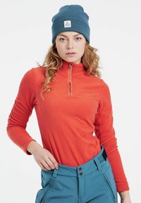 Fleeceová mikina v jasne oranžovej farbe s predným zipsom a dlhými rukávmi. Skombinovaná s modrými lyžiarskymi nohavicami s dvoma gombíkmi a pútkami na opasok.