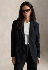 Žena s dugom kovrčavom kosom, nosi crne sunčane naočale, crni sako, bijelu majicu i crne hlače, na jednostavnoj bež pozadini.