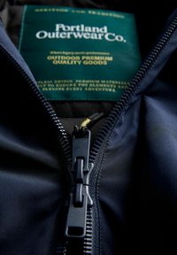 Jūras spēku žakete ar melnu rāvējslēdzēju, kurai ir teksturēts audums. Uz etiķetes rakstīts "Portland Outerwear Co." baltā krāsā, uzsverot augstas kvalitātes kvalitatīvos āra apģērbus.