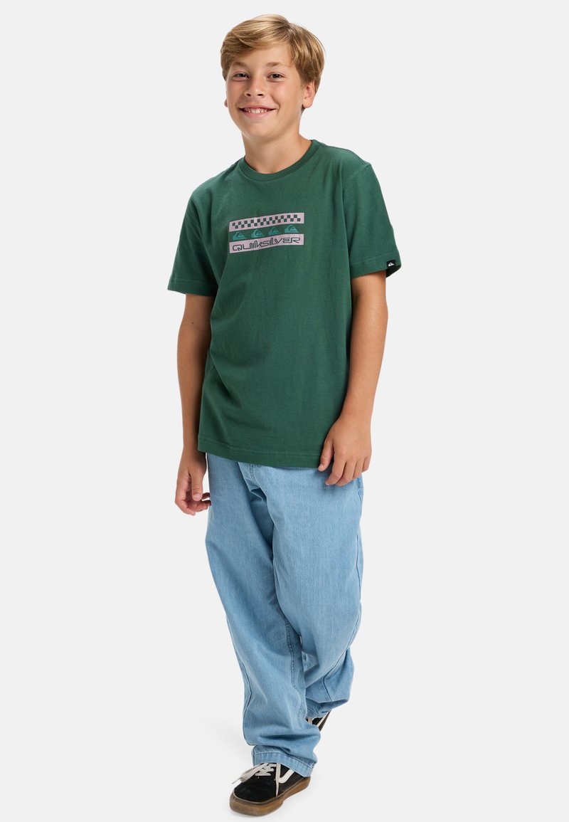 Zielona koszulka bawełniana z graficznym nadrukiem i krótkimi rękawami, w połączeniu z jasnoniebieskimi dżinsami i czarnymi trampkami.