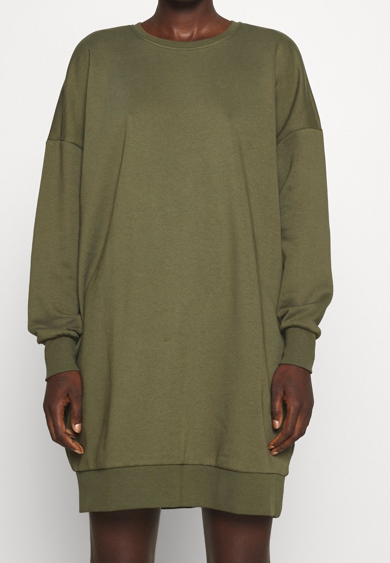 Personne portant une robe-sweat oversize à manches longues vert olive avec poignets et ourlet côtelés, debout devant un fond blanc uni.