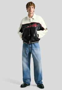 Veste varsity en cuir noir et blanc avec lettrage rouge, pantalon en denim bleu à coupe décontractée et chaussures noires, présentés sur un fond neutre.