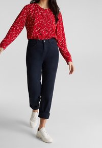 Femme portant un blouse à fleurs rouge à manches longues, un pantalon navy retroussé et des baskets blanches marchant devant un fond uni.