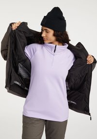 Svetlofialový pulóver na zips do polovice s jemnou textúrou, nosený pod tmavým, zatepleným kabátom. Má vysoký golier a vrecká.