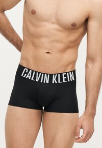 Mandlig overkrop iført sorte Calvin Klein boxer briefs med synligt logo på linningen, beskåret ved midt-låret og øverste bryst.