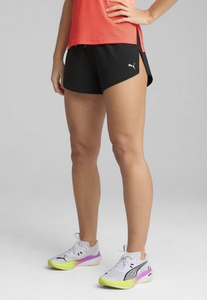 Schwarze Sportshorts mit einem schwarzen Puma-Logo, kombiniert mit weißen und Neon-Schuhen mit lila Akzenten. Das Oberteil hat eine leuchtend rote Netzstruktur.
