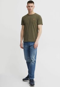 Olivgrünes T-Shirt mit kurzen Ärmeln, dezenten Streifen und kleinem roten Logo, kombiniert mit hellblauen Jeans und dunklen Sneakern.