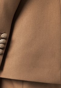 Beige skräddarsydd kostymärm med fyra matchande knappar täckta av tyg i rad, som visar en fin twillvävstextur.