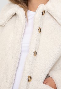 Weiße Kunstpelzjacke mit weicher Textur, großem Kragen und fünf runden Knöpfen in Messingfarbe. Die innere Schicht besteht aus einem schlichten weißen Hemd.