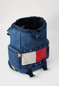 Sac à dos en denim avec cordon de serrage bleu marine, panneau en tissu rouge et blanc contrasté, poche zippée à l'avant et bretelles réglables.