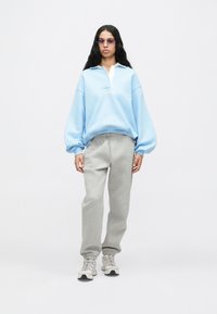 Žena v svetlomodrej oversized mikine, sivých teplákových nohaviciach, teniskách a fialových okuliaroch stojí na pozadí jednoduchej bielej steny.