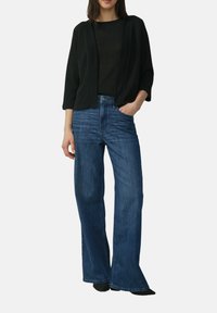 Zwarte open voorkardigan over een donkere top, gecombineerd met uitlopend blauwe spijkerbroeken. De materialen lijken zacht; de denim heeft een klassieke wassing.