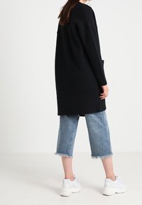 Musta pitkä neuletakki, jossa on teksturoidun kankaan pinta, väljä istuvuus ja taskut. Yhdistetty vaaleansinisiin lyhytlahkeisiin farkkuihin ja valkoisiin tennareihin.