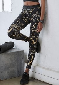 Black leggings with a gold marbled pattern, stretch fabric, a high waistband, and ankle length. Worn with black athletic shoes.