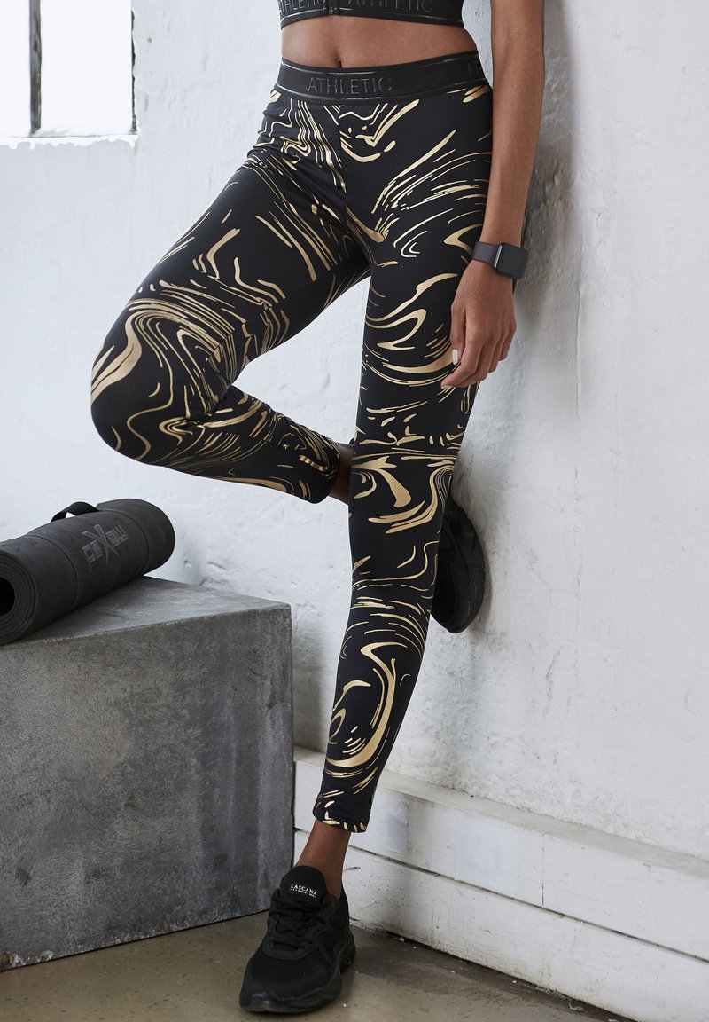 Black leggings with a gold marbled pattern, stretch fabric, a high waistband, and ankle length. Worn with black athletic shoes.
