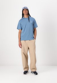 T-shirt bleu clair à manches courtes, pantalon beige ample, chaussures de sport navy et une casquette bleu clair assortie avec un logo. Design simple et décontracté.
