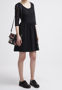 Schwarzes Spitzenkleid mit dreißig Zentimeter langen Ärmeln, tailliertem Oberteil und ausgestelltem Rock. Kombiniert mit einer gemusterten Handtasche und schwarzen Schuhen.